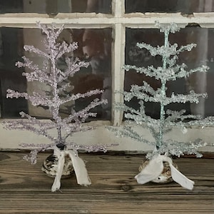 May include: Two small, decorative Christmas trees. One is light purple, the other is silver. Both have a textured, crystallized appearance and are adorned with white ribbon. They sit on a wooden surface in front of a window.