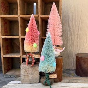 May include: Three decorative bottle brush trees in shades of pink and teal, adorned with pastel-colored Easter egg ornaments. One tree has a tag that says "spring", and another says "easter". The trees are displayed on rustic wooden and metal bases.