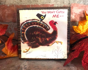 May include: A wooden sign with a vintage illustration of a turkey wearing a pilgrim hat and running. The text on the sign reads "You won't catch me--".