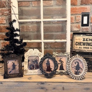 May include: Five decorative picture frames in various shapes and finishes, each containing a silhouette illustration of a witch. The frames feature ornate, detailed borders in black, white, and metallic tones, displaying imagery of witches with brooms, cauldrons, and pointed hats.
