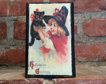 May include: A vintage Halloween greeting card featuring a girl in a witch's hat holding a black cat. The card has the text "Hallowe'en Greetings."