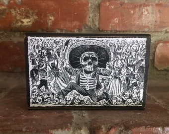 May include: Black and white wood block print of a skeleton wearing a sombrero and holding a scythe. The skeleton is surrounded by other skeletons and skulls. The print is on a black background.