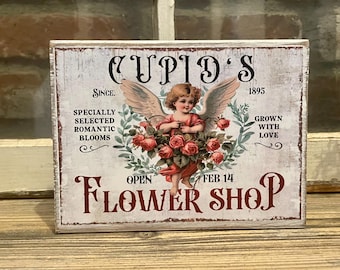 May include: A white wooden sign with the text "Cupid's Flower Shop" in red lettering. The sign features a vintage illustration of a cherub holding a bouquet of red roses. The text "Since 1895" and "Grown with Love" are also on the sign.