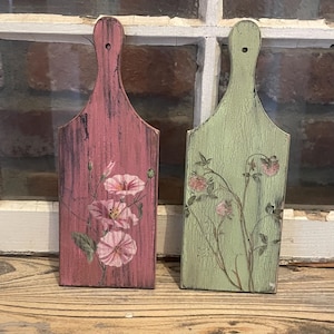 May include: Two decorative wooden cutting boards. One is painted pink with a floral design, the other is green with a floral design. Both have a handle with a hole for hanging. The boards have a distressed, vintage look.