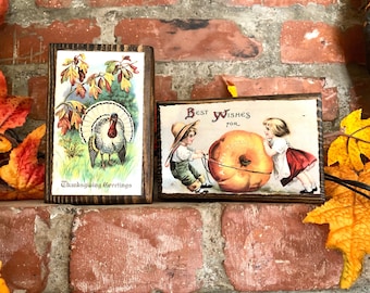 May include: Two vintage wooden plaques with Thanksgiving-themed illustrations. The first plaque features a turkey with the text "Thanksgiving Greetings". The second plaque features two children pushing a large pumpkin with the text "Best Wishes For".