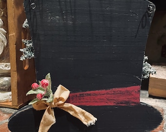 May include: Black wooden top hat decoration with a red band, adorned with a beige ribbon and faux berries. The hat has a distressed finish and metal wire hangers for display. A festive, rustic holiday decoration.