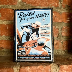 May include: A vintage wooden sign with a blue and white illustration of a sailor working on a ship. The sign reads "Build for your NAVY!" and encourages enlistment in the Navy. It also lists the occupations needed, such as carpenters, machinists, and electricians. The sign is on a brick background.