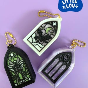 May include: Three gothic window-shaped keychains with gold ball chains. One is black with a green butterfly design, one is white with a black bat and moon, and one is iridescent purple with a black frame. Each features intricate window pane details.