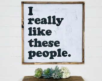 May include: A white wooden sign with a brown frame, hanging on a white wall. The sign says "I really like these people." in black bold letters.