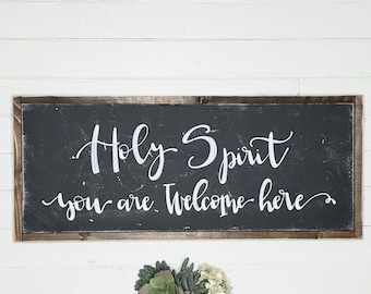 May include: A black wooden sign with a white inscription that reads "Holy Spirit you are welcome here". The sign is framed with a dark wood frame and has a distressed look.