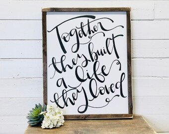 May include: A white wooden sign with black calligraphy that reads "Together they built a life they loved".