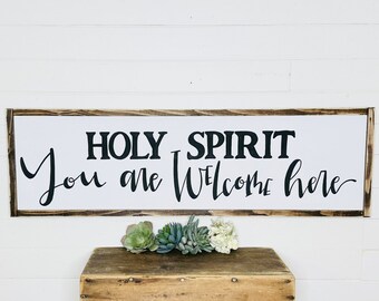 May include: A white wooden sign with a black wood frame and the text "HOLY SPIRIT You are welcome here" in black lettering. The sign is hanging on a white wall with a small wooden box with green succulents in front of it.
