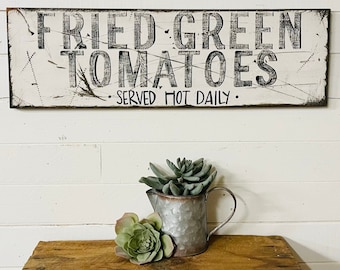 May include: White wooden sign with black lettering that reads "Fried Green Tomatoes - Served Hot Daily". The sign is hanging on a white wall. A small metal watering can with a succulent plant is sitting on a wooden table.