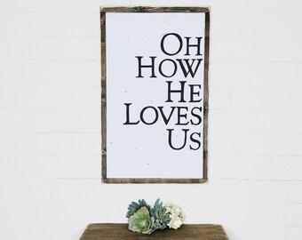 May include: A rectangular wooden sign with the words "OH HOW HE LOVES US" in black lettering. The sign has a white background and a dark brown wooden frame. A small arrangement of succulents and white flowers sits on a wooden surface below the sign.