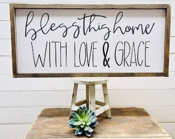 May include: A wooden sign with a white background and black lettering that reads "bless this home with love & grace".