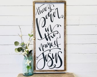 May include: A wooden sign with a white background and black calligraphy that reads "There is power in the name of Jesus".
