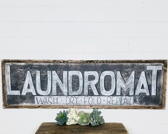 May include: A rustic wooden sign with a black background and white lettering that reads "Laundromat" and "Wash - Dry - Fold - Repeat".