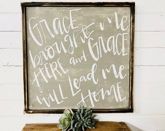 May include: A framed sign with a beige background and white lettering that reads "Grace brought me here and Grace will lead me home".