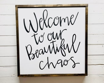 May include: A white wooden sign with a brown frame, featuring black calligraphy that reads "Welcome to our beautiful chaos".