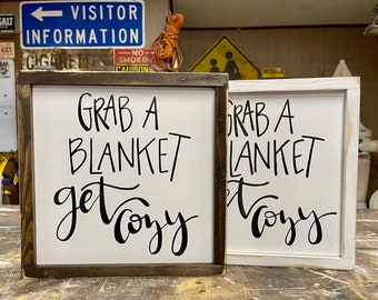 May include: Two wooden signs with white backgrounds and black lettering. The signs say "Grab a blanket get cozy".