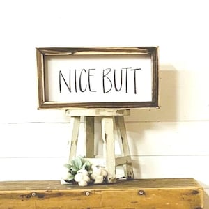 May include: A rustic wooden sign with the text "NICE BUTT" in black lettering. The sign is displayed on a small white stool with a green plant.