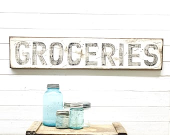 May include: A white wooden sign with black lettering that reads "GROCERIES". The sign is hanging on a white wood panel wall. There are four blue glass jars on a wooden surface in front of the sign.