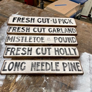 Może przedstawiać: Pięć drewnianych tabliczek z czarnym napisem na białym tle. Tabliczki głosi: "Fresh Cut-U Pick", "Fresh Cut Garland", "Mistletoe by the Pound", "Fresh Cut Holly", i "Long Needle Pine".