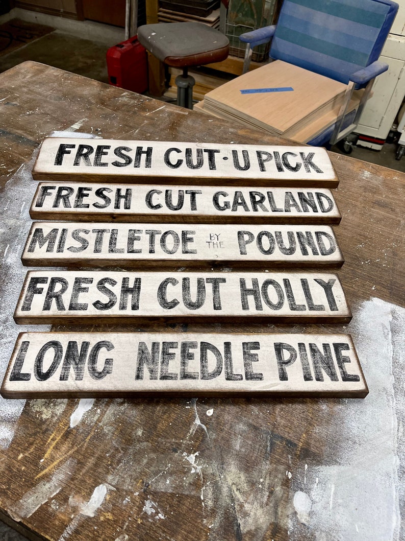 Może przedstawiać: Pięć drewnianych tabliczek z czarnym napisem na białym tle. Tabliczki głosi: "Fresh Cut-U Pick", "Fresh Cut Garland", "Mistletoe by the Pound", "Fresh Cut Holly", i "Long Needle Pine".