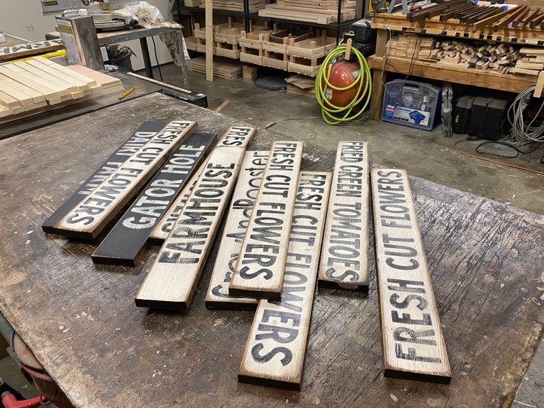 Może przedstawiać: Kolekcja drewnianych tabliczek z r&oacute;żnymi napisami, w tym "Fresh Cut Flowers", "Farmhouse", "Gator Hole" i "Fried Green Tomatoes". Tabliczki są ułożone na stole w warsztacie.