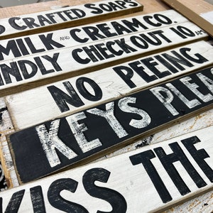 Może przedstawiać: Pięć drewnianych tabliczek z czarnymi napisami na białym tle. Tabliczki głosi: "Crafted Soaps", "Milk and Cream Co.", "Kindly Check Out 10 AM", "No Peeing", "Keys Please", i "Kiss The".