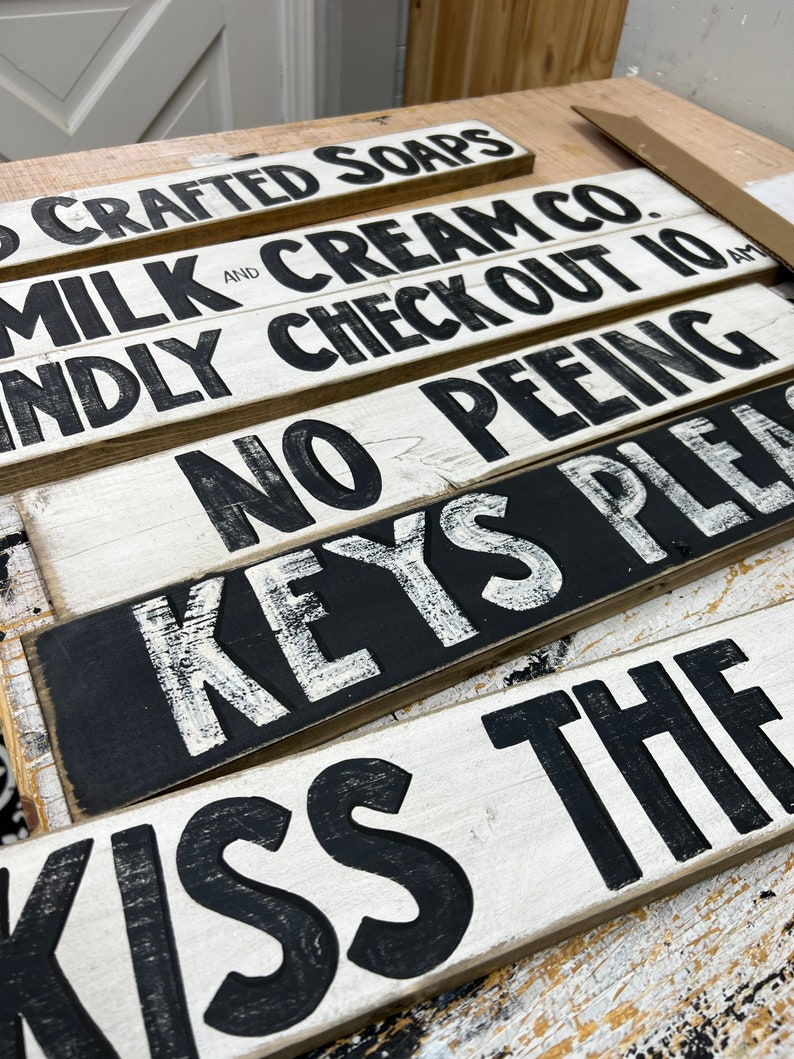 Może przedstawiać: Pięć drewnianych tabliczek z czarnymi napisami na białym tle. Tabliczki głosi: "Crafted Soaps", "Milk and Cream Co.", "Kindly Check Out 10 AM", "No Peeing", "Keys Please", i "Kiss The".