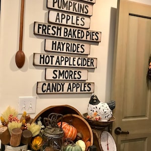 Może przedstawiać: Drewniany znak z napisem "FRESH PICKED", po kt&oacute;rym następuje seria znak&oacute;w z jesiennymi napisami, takimi jak "SUNFLOWERS", "PUMPKINS", "APPLES", "FRESH BAKED PIES", "HAYRIDES", "HOT APPLE CIDER", "SMORES" i "CANDY APPLES". Znaki wiszą na białej ścianie. Jest drewniana miska z kilkoma dyniami i dyniami, szklany słoik z lampkami i małe drewniane pudełko z napisem "FARMERS MARKET".