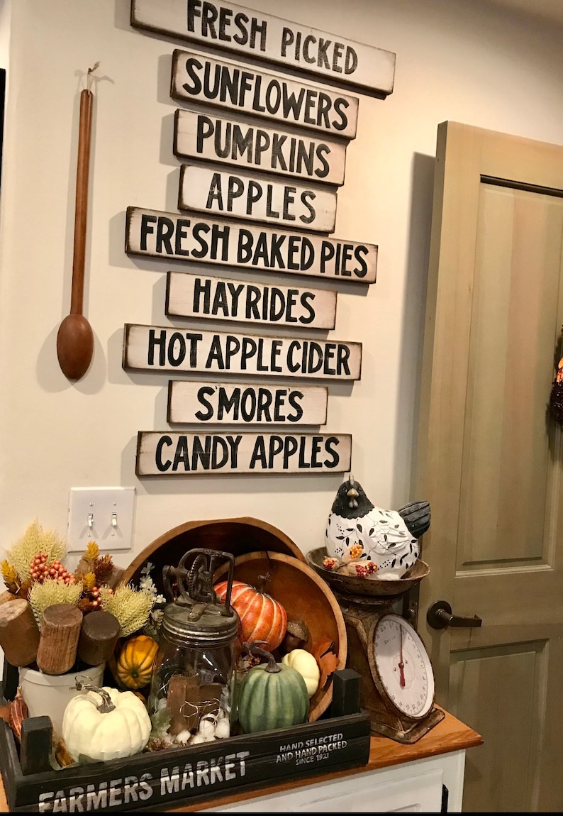 Może przedstawiać: Drewniany znak z napisem "FRESH PICKED", po kt&oacute;rym następuje seria znak&oacute;w z jesiennymi napisami, takimi jak "SUNFLOWERS", "PUMPKINS", "APPLES", "FRESH BAKED PIES", "HAYRIDES", "HOT APPLE CIDER", "SMORES" i "CANDY APPLES". Znaki wiszą na białej ścianie. Jest drewniana miska z kilkoma dyniami i dyniami, szklany słoik z lampkami i małe drewniane pudełko z napisem "FARMERS MARKET".