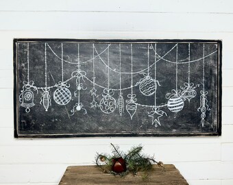 May include: A rectangular chalkboard with a black frame features a white chalk drawing of Christmas ornaments hanging from strings. The ornaments include bells, baubles, stars, and teardrop shapes. The chalkboard is mounted on a white wooden wall.