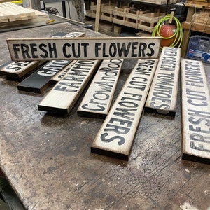 Może przedstawiać: Zbi&oacute;r drewnianych tabliczek z czarnymi krawędziami i białymi napisami. Tabliczki głosi: "FRESH CUT FLOWERS", "FRESH CUT FLOWERS", "CROW AND ROD", "FARMHOUSE", "FRESH GREEN TOMATOES" i "GAT" .