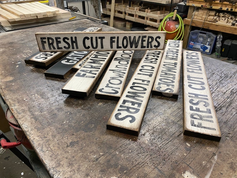 Może przedstawiać: Zbi&oacute;r drewnianych tabliczek z czarnymi krawędziami i białymi napisami. Tabliczki głosi: "FRESH CUT FLOWERS", "FRESH CUT FLOWERS", "CROW AND ROD", "FARMHOUSE", "FRESH GREEN TOMATOES" i "GAT" .