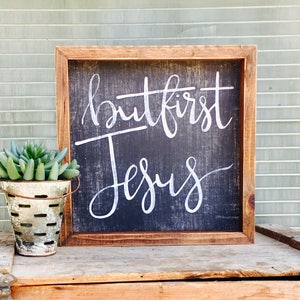 May include: A wooden framed sign with a black background and white lettering that reads "but first Jesus". A small galvanized metal bucket with a succulent plant is in the foreground.