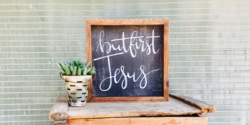 May include: A wooden framed sign with a black background and white lettering that reads "but first Jesus". A small galvanized metal bucket with a succulent plant is in the foreground.