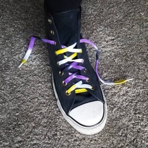 Pode incluir: Tênis preto de cano alto com biqueira branca e cadarços coloridos. Os cadarços são uma mistura de amarelo, branco e roxo. O tênis está em um tapete cinza.