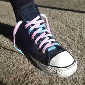 May include: A navy blue high-top trainer with a white rubber toe cap and sole. The laces are a gradient of pink, blue, and white. The shoe is on a neutral-coloured carpet.