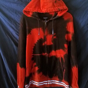 Puede incluir: Sudadera con capucha roja y negra tie-dye con rayas blancas en la parte inferior. La sudadera tiene capucha y un bolsillo delantero. El nombre de la marca "ARIZONA" est&aacute; impreso en la parte delantera de la sudadera.