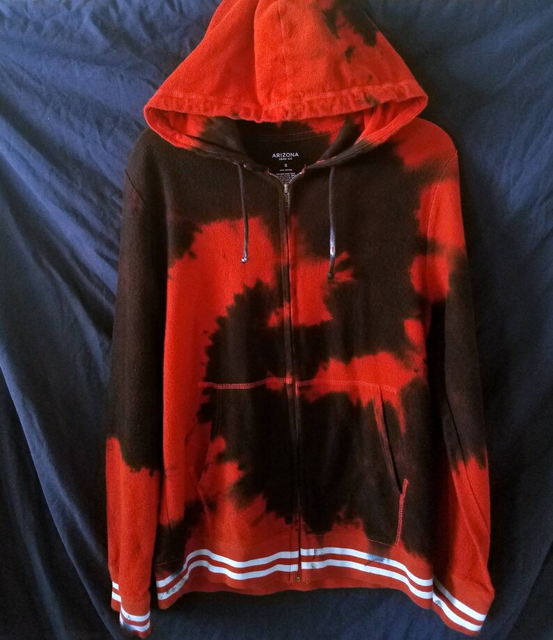 Puede incluir: Sudadera con capucha roja y negra tie-dye con rayas blancas en la parte inferior. La sudadera tiene capucha y un bolsillo delantero. El nombre de la marca "ARIZONA" est&aacute; impreso en la parte delantera de la sudadera.