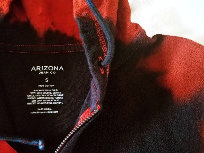 Puede incluir: Una sudadera con capucha de color negro y rojo con efecto tie-dye y cierre de cremallera. La etiqueta dice "Arizona Jean Co" y "S" para el tama&ntilde;o. Las instrucciones de cuidado incluyen lavar a m&aacute;quina con agua fr&iacute;a, ciclo delicado, usar solo blanqueador sin cloro si es necesario, secar en secadora a baja temperatura, planchar a temperatura media si es necesario, no planchar sobre la impresi&oacute;n. Hecho en India. Proveedor 76441-5RNP93677.