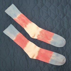 Puede incluir: Un par de calcetines teñidos con un puño y puntera azules, un centro rosa y una sección media blanca. Los calcetines se muestran sobre una superficie acolchada azul oscuro. Los calcetines están hechos de un material suave y cómodo.