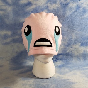 May include: Pink fleece hat with a crying face emoji design. The eyes are black with white pupils, the mouth is black, and the tears are light blue.