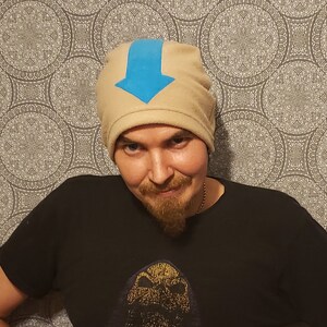 May include: A person wearing a tan beanie with a blue arrow on the front. The beanie is made of fleece and has a fold-over cuff.