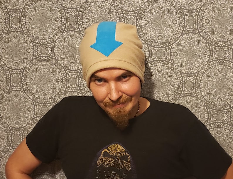 May include: A person wearing a tan beanie with a blue arrow on the front. The beanie is made of fleece and has a fold-over cuff.