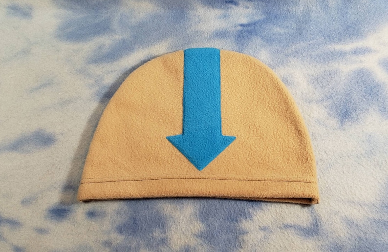 May include: A beige fleece beanie with a blue arrow pointing down on the front.