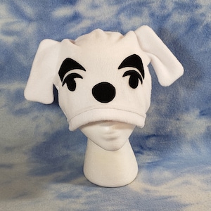 May include: White fleece hat with black felt appliques of a dog's face, including eyes, nose, and eyebrows. The hat has two floppy ears.