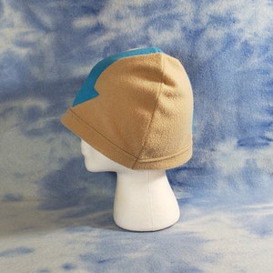 May include: A tan fleece beanie with a blue arrow design on the front. The beanie is on a white mannequin head.
